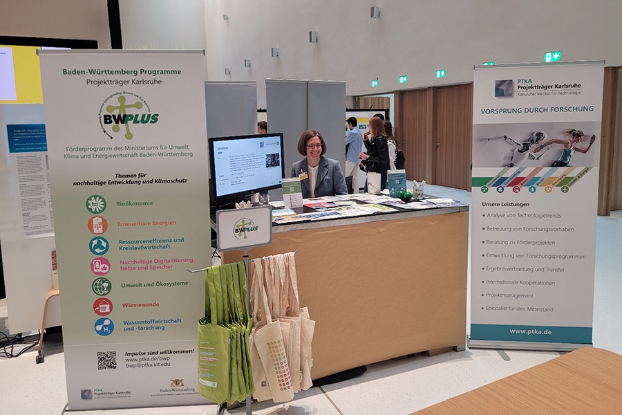 Eine Mitarbeiterin am Messestand des PTKA beim 13. Ressourceneffizienz- und Kreislaufwirtschaftskongress Baden-Württemberg mit Infomaterial und Roll-Ups freut sich darauf, Interessierte zu beraten.